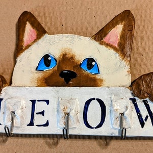 Puede incluir: Llavero de madera pintado a mano con una cara de gato con ojos azules y la palabra "MEOW" debajo. El gato es de color crema y marrón. Incluye cuatro ganchos metálicos para llaves.