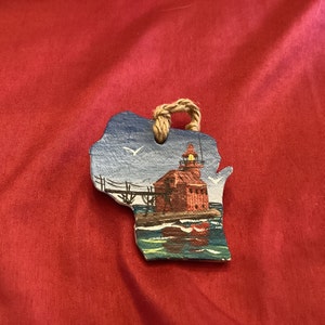 May include: A wooden ornament shaped like the state of Wisconsin with a hand-painted image of a red lighthouse on a blue body of water. The ornament has a rope hanger.
