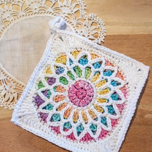 May include: A square, white crocheted potholder with a colorful floral design in the center. The flower pattern features shades of pink, yellow, green, and blue. A white crocheted loop is attached for hanging. A white lace doily is in the background.
