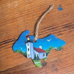 May include: A hand-painted wooden ornament in the shape of a state, featuring a lighthouse and a house with a red roof. The ornament has a blue sky background with white seagulls and a jute string for hanging.