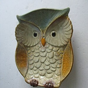 May include: Ceramic owl-shaped dish with a textured surface. The owl has large, black eyes, a yellow beak, and a patterned body in shades of beige, brown, and green. The dish is designed to resemble an owl.