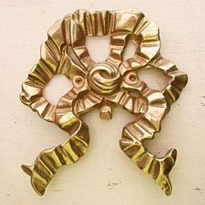 May include: A gold-toned metal decorative piece with a unique, intricate design resembling a stylized flower or ribbon. The piece has a textured, ornate surface and a slightly raised center.