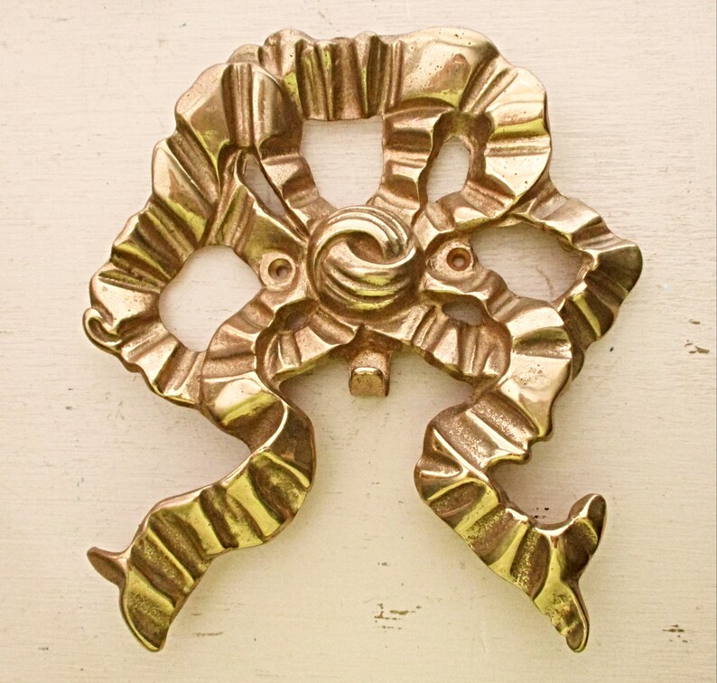 May include: A gold-toned metal decorative piece with a unique, intricate design resembling a stylized flower or ribbon. The piece has a textured, ornate surface and a slightly raised center.