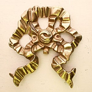 May include: A gold-colored metal decorative bow with a detailed, textured design. The bow is made of a single piece of metal and has a slightly raised center.