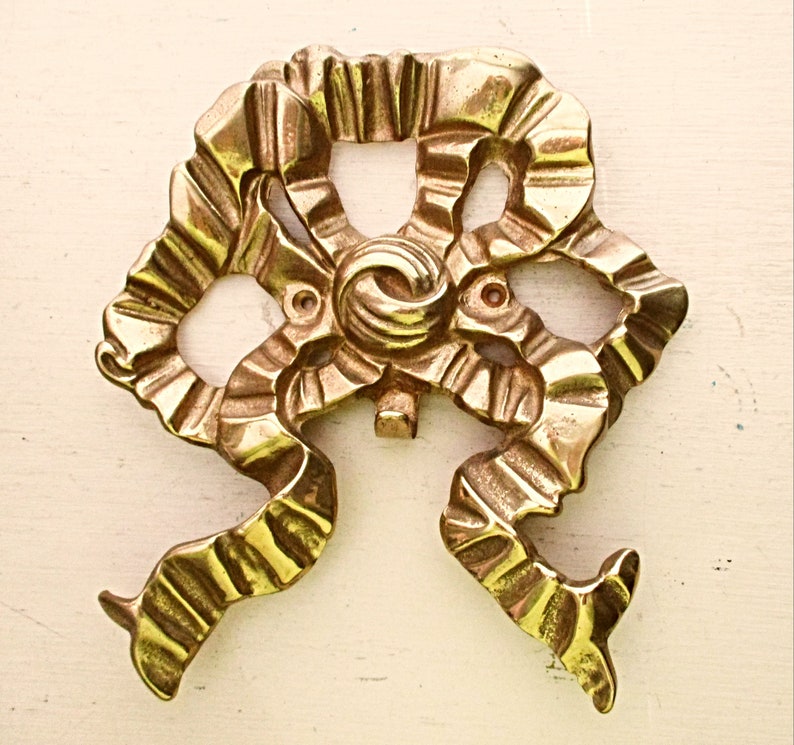 May include: A gold-colored metal decorative bow with a detailed, textured design. The bow is made of a single piece of metal and has a slightly raised center.