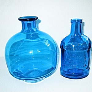 May include: Two blue glass bottles. One is a round, bulbous shape with a narrow neck. The other is a smaller, rectangular bottle with a rounded top. Both bottles are a vibrant shade of blue and appear to be empty.