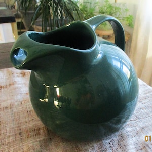 May include: A dark green ceramic pitcher with a rounded body and a handle. The pitcher has a smooth, glossy finish.