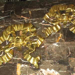 May include: Two gold-colored metal wall sconces with a decorative, ornate design. The sconces are shaped like a ribbon or bow and have a textured, wavy surface.