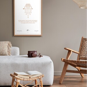 May include: A framed print with a gold design and text, hanging on a neutral wall. The text reads, "In this moment, I meet myself with kindness. It is safe for my thoughts to slow and soften. I am supported as I find my calm." A brown leather camera case sits on a white ottoman.