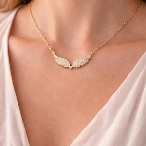 Puede incluir: Un collar dorado con un par de colgantes de alas. Cada ala est&aacute; incrustada con peque&ntilde;as piedras brillantes. El collar lo lleva una persona que viste una blusa blanca.
