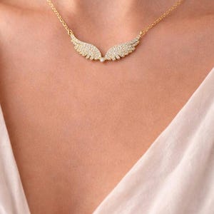 Puede incluir: Un collar dorado con un colgante en forma de dos alas. Las alas est&aacute;n incrustadas con peque&ntilde;os cristales brillantes. El collar se lleva en el pecho de una persona, sobre una tela clara.