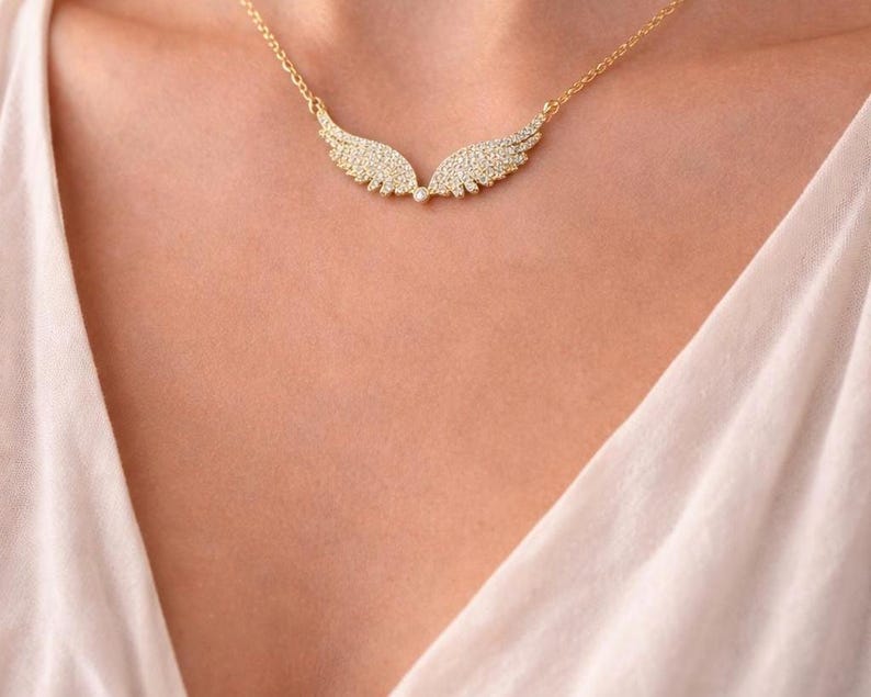 Puede incluir: Un collar dorado con un colgante en forma de dos alas. Las alas est&aacute;n incrustadas con peque&ntilde;os cristales brillantes. El collar se lleva en el pecho de una persona, sobre una tela clara.