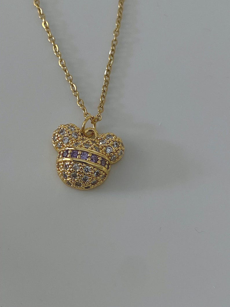 May include: A gold chain necklace with a small, sparkly Mickey Mouse pendant. The pendant is covered in clear and purple rhinestones.