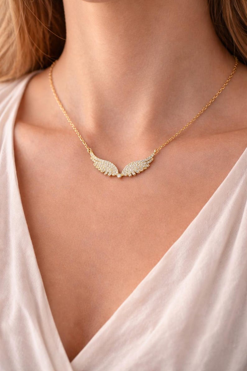 Puede incluir: Un collar dorado con un colgante en forma de alas. Cada ala est&aacute; adornada con peque&ntilde;as piedras brillantes. El collar se muestra en una persona que lleva una camiseta blanca.