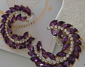 May include: A pair of gold earrings with a purple and clear crystal design. The earrings are shaped like a crescent moon and are adorned with small, sparkling crystals.
