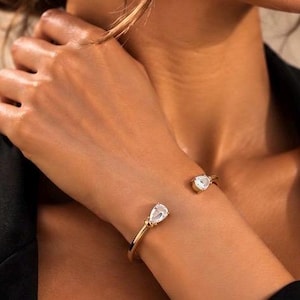 Könnte beinhalten: Ein goldfarbenes, offenes Armband mit zwei tropfenförmigen, klaren Steinen an den Enden. Das Armband besteht aus einem glatten, polierten Metallband, das vorne offen ist und zwei facettierte Kristalle als Blickfang präsentiert.
