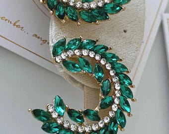May include: A pair of gold-tone earrings featuring a crescent-shaped design adorned with marquise-cut emerald green crystals and a row of small, round, clear white rhinestones. The earrings are attached to a white display card with partial text.