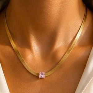 Op de afbeelding: Een goudkleurige visgraatketting met een enkele vierkant geslepen lichtroze edelsteen als hanger in het midden. De platte, flexibele metalen schakels vormen een gladde, doorlopende band die mooi rond de hals valt en de steen accentueert.