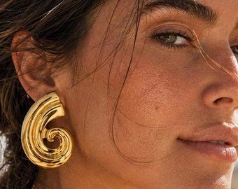 May include: A large, polished gold-tone earring featuring a sculptural, swirling shell-like design. The earring has a smooth, reflective metallic surface with a prominent spiral shape that curves inward, creating a bold and dimensional appearance.