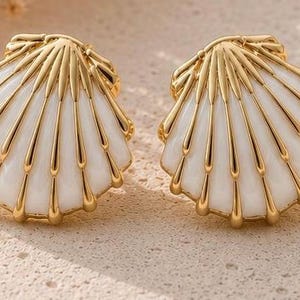 May include: A pair of earrings shaped like scallop shells, featuring a glossy white enamel finish with raised, polished gold-tone metal ridges radiating from the top hinge to the scalloped bottom edge, creating a textured, fan-like appearance.