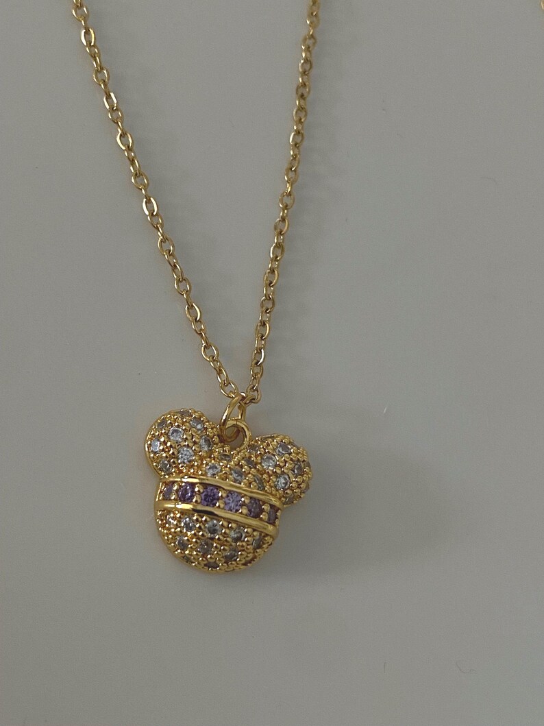 May include: A gold chain necklace with a small, sparkly Mickey Mouse pendant. The pendant is covered in clear and purple rhinestones.