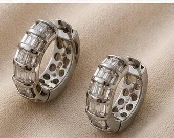 May include: A pair of silver-toned hoop earrings featuring a row of clear, rectangular baguette-cut stones set along the outer curve. The inner side of the hoops displays a series of circular cutouts, and the metal has a polished, reflective finish.