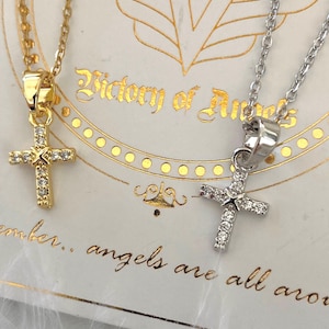 Op de afbeelding: Twee delicate kettingen met kruis hangers. Het kruis aan de linkerkant is goud met heldere stenen, en het kruis aan de rechterkant is zilver met heldere stenen. De kettingen worden getoond op een witte kaart met de tekst "Victory of Angels" en "Remember... angels are all around".
