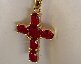 May include: Gold-colored necklace featuring a cross pendant. The cross is adorned with oval-shaped, red gemstones. The chain and pendant are a matching gold color, creating a cohesive and elegant design. The necklace is displayed against a neutral background.