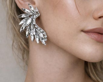 May include: A silver-toned ear crawler earring featuring a cluster of clear, marquise-cut crystals arranged in a wing-like shape, accented with smaller round crystals along the inner curve that follows the contour of the earlobe.