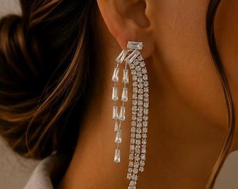 May include: A long, silver-toned dangle earring featuring a rectangular stud at the top. The design includes two distinct sections: one side with three vertical strands of round clear crystals and the other with three shorter strands of baguette-cut clear crystals.