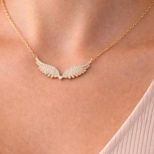Puede incluir: Un collar dorado con un colgante en forma de alas de &aacute;ngel. Las alas est&aacute;n adornadas con peque&ntilde;as piedras brillantes. El collar se muestra en el pecho de una persona.