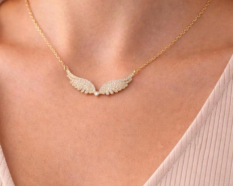 Puede incluir: Un collar dorado con un colgante en forma de alas de &aacute;ngel. Las alas est&aacute;n adornadas con peque&ntilde;as piedras brillantes. El collar se muestra en el pecho de una persona.