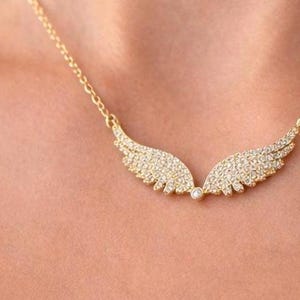 Puede incluir: Un collar dorado con un dise&ntilde;o de alas. Cada ala est&aacute; incrustada con peque&ntilde;as piedras preciosas transparentes. El collar se muestra sobre la piel de una persona.