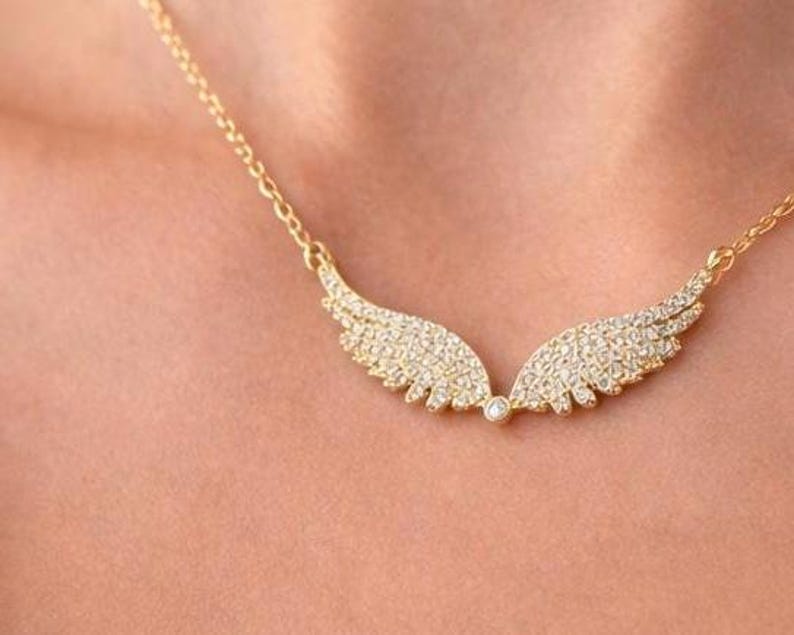 Puede incluir: Un collar dorado con un dise&ntilde;o de alas. Cada ala est&aacute; incrustada con peque&ntilde;as piedras preciosas transparentes. El collar se muestra sobre la piel de una persona.
