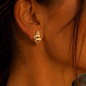 Könnte beinhalten: Ein goldener Ohrstecker in Form einer kleinen, spiralförmigen Muschel mit strukturierter Oberfläche. Der Ohrstecker wird am Ohrläppchen getragen, hat ein metallisches Finish und eine spitz zulaufende Unterseite, die das Licht auf der Struktur reflektiert.