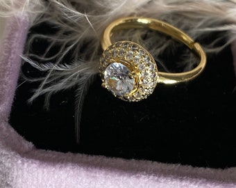 May include: A gold ring with a large, clear, round gemstone surrounded by smaller, clear gemstones. The ring is in a small, purple velvet box.