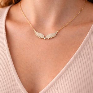 Puede incluir: Un collar dorado con un colgante en forma de alas de &aacute;ngel, adornado con peque&ntilde;os cristales brillantes. El collar lo lleva una persona con una camiseta rosa claro. Las alas est&aacute;n detalladas.