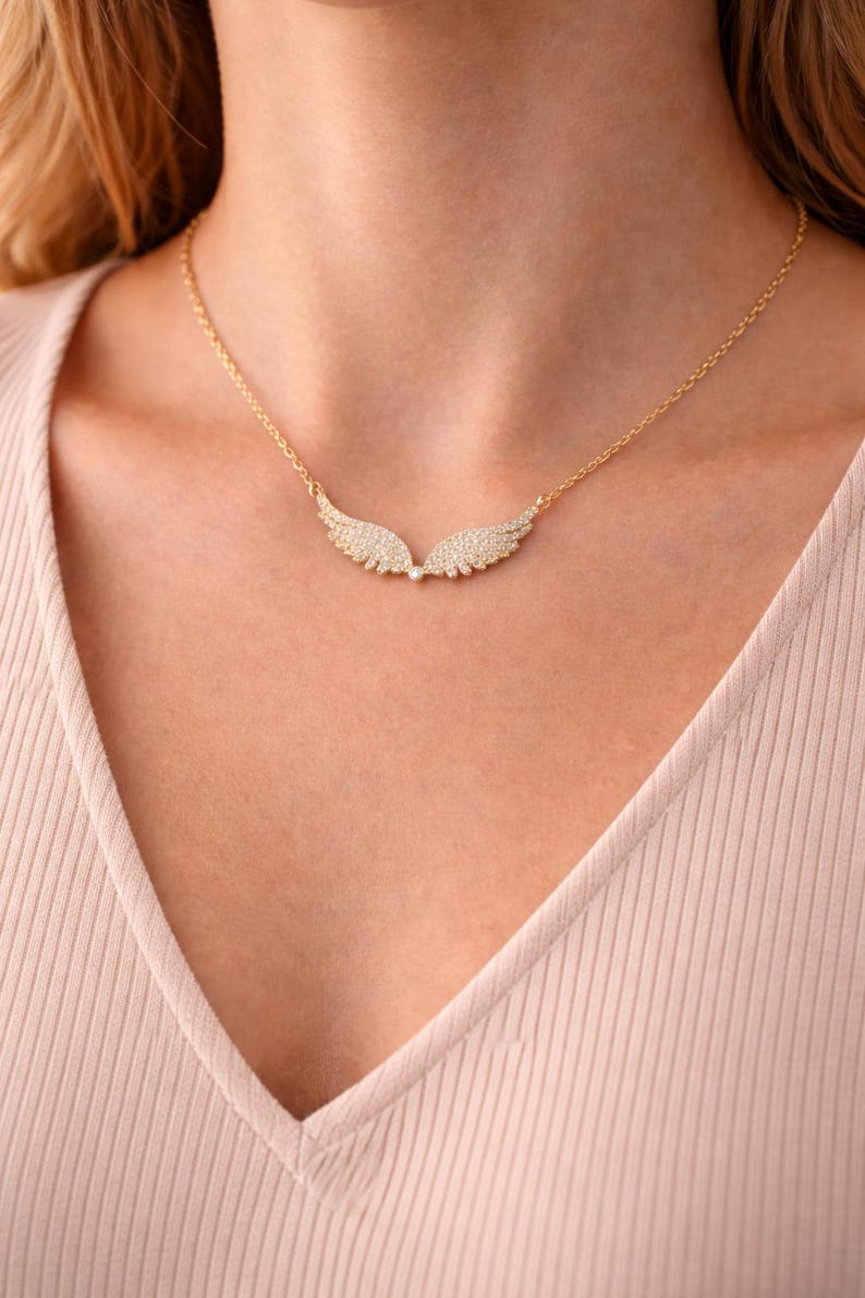 Puede incluir: Un collar dorado con un colgante en forma de alas de &aacute;ngel, adornado con peque&ntilde;os cristales brillantes. El collar lo lleva una persona con una camiseta rosa claro. Las alas est&aacute;n detalladas.