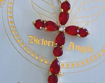 Op de afbeelding: Een goudkleurige kruishanger aan een ketting met meerdere rode peervormige en ronde geslepen stenen in een kruispatroon en een heldere steen in het midden. De hanger ligt op een witte kaart met de gouden tekst Victory Angels.