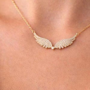 Puede incluir: Un collar dorado con un colgante en forma de alas de &aacute;ngel. Las alas est&aacute;n incrustadas con peque&ntilde;as piedras brillantes. El collar se lleva alrededor del cuello. Una delicada pieza de joyer&iacute;a.