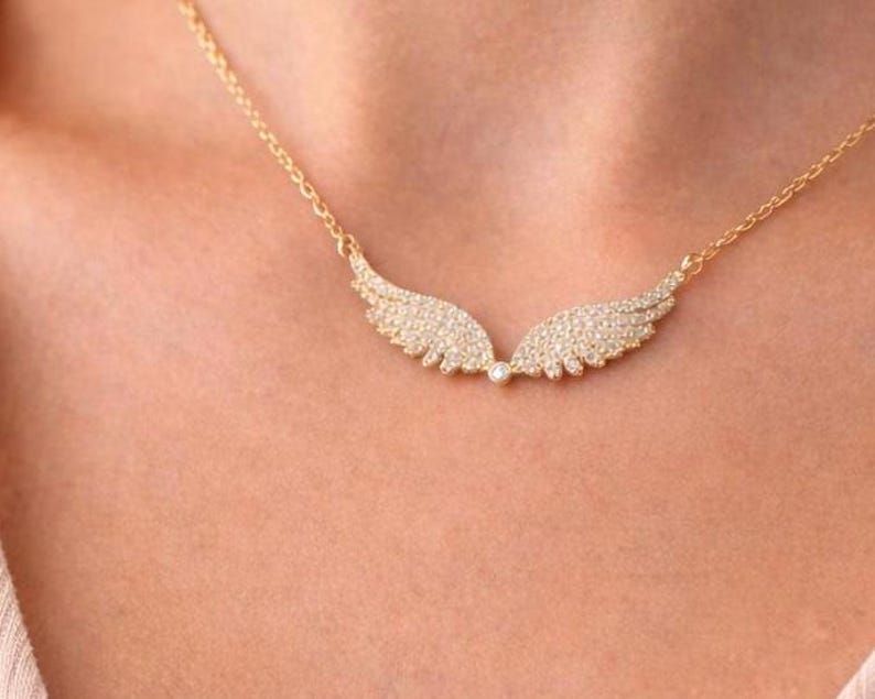 Puede incluir: Un collar dorado con un colgante en forma de alas de &aacute;ngel. Las alas est&aacute;n incrustadas con peque&ntilde;as piedras brillantes. El collar se lleva alrededor del cuello. Una delicada pieza de joyer&iacute;a.