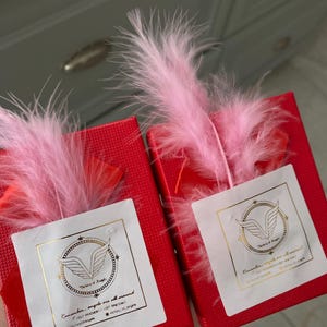 May include: Two red gift boxes with white labels and gold lettering. The labels feature a gold angel wing logo and the text "Remember, angels are all around." Each box is topped with a pink feather.