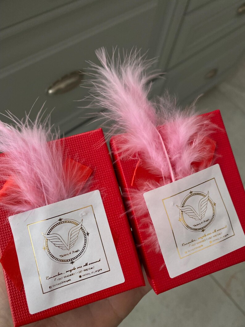 May include: Two red gift boxes with white labels and gold lettering. The labels feature a gold angel wing logo and the text "Remember, angels are all around." Each box is topped with a pink feather.