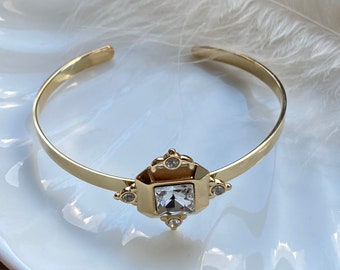 May include: A gold bangle bracelet with a clear square crystal in the center. The crystal is surrounded by smaller gold accents and clear stones.