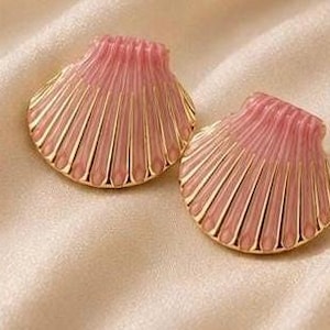 May include: A pair of shell-shaped earrings featuring a soft pink translucent center with raised gold-toned metallic ridges radiating from the top hinge. The design mimics the appearance of a scallop shell with a polished finish and gold edges.