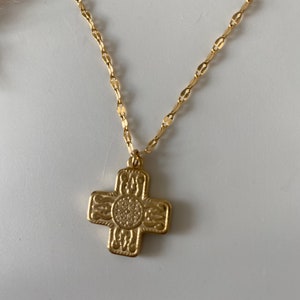 May include: A gold-colored necklace with a cross pendant. The cross has an ornate design with a circular center and decorative patterns on each arm. The chain is a delicate, linked style.