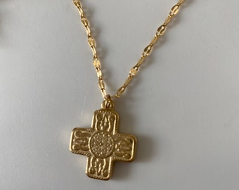 May include: A gold-colored necklace with a cross pendant. The cross has an ornate design with a circular center and decorative patterns on each arm. The chain is a delicate, linked style.