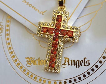 Op de afbeelding: Een goudkleurige ketting met kruishanger, voorzien van een centrale rij feloranje ronde stenen omrand door kleine heldere witte kristallen. De hanger hangt aan een bijpassende goudkleurige ketting op een witte kaart met de tekst Victoria Angels.