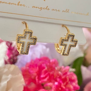 Puede incluir: Pendientes de cruz dorados con centros de esmalte blanco y detalles brillantes. Los pendientes se muestran sobre un fondo de flores rosas y blancas, con el texto "remember... angels are all around" visible.