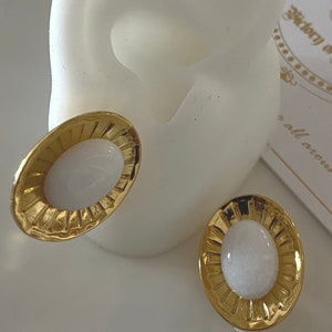 Puede incluir: Un par de pendientes de clip ovalados de oro con piedras cabujón blancas en el centro. Los pendientes tienen un diseño texturizado en forma de sol alrededor de las piedras.